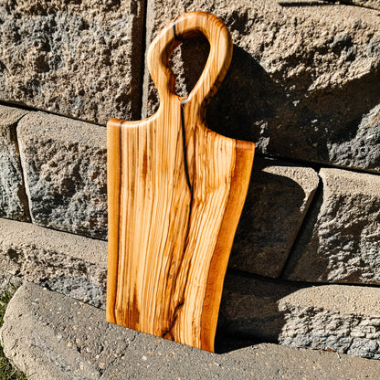 A rustic live-edge wooden cutting board with a loop handle, leaning against a stone wall outdoors.